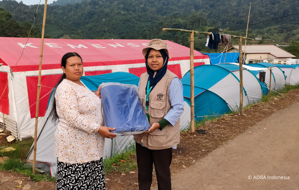 eine ADRA-Mitarbeiterin verteilt Planen und Baumaterialien an Menschen in einem von einem Erdbeben betroffenen Gebiet in Cianjur, Indonesien. Im Hintergrund sind aufgebaute Notunterkünfte in Form von Zelten zu sehen.