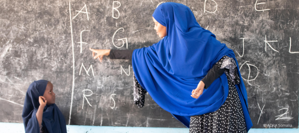 Engagierte Lehrerin in Somalia zeigt auf den Buchstaben 'F' an einer Tafel, während ein aufgewecktes Schulmädchen neben ihr steht