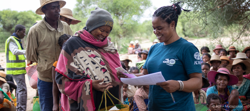 ADRA-Mitarbeiterin übergibt einer Frau in Madagaskar Unterlagen, im Hintergrund warten Menschen auf Hilfsgüter