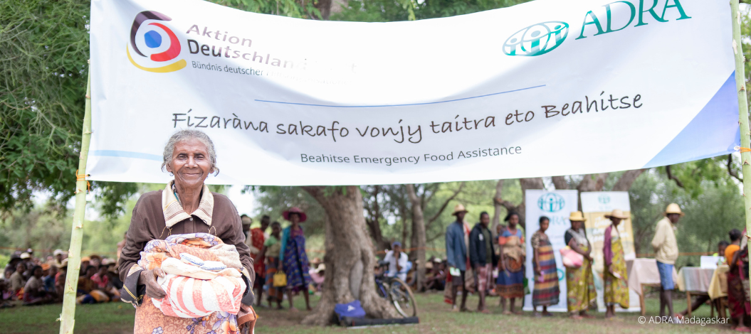 Ältere Frau in Madagaskar hält Hilfsgüter vor ADRA- und ADH-Banner, hinten warten Menschen in der Schlange
