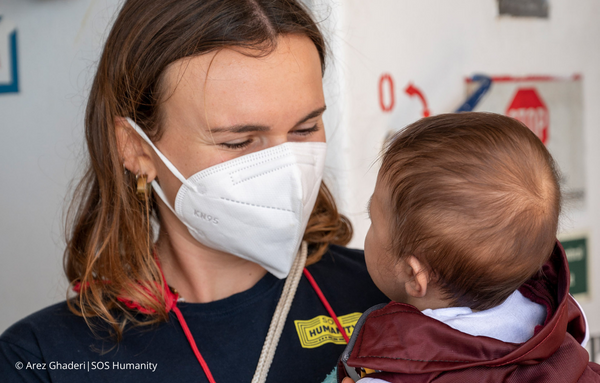 Eine Helferin von SOS Humanity trägt eine Atemschutzmaske und hält liebevoll ein gerettetes Kind in ihren Armen. Trotz der Maske ist zu erkennen, dass sie das Kind anlächelt. © Arez Ghaderi_SOS Humanity