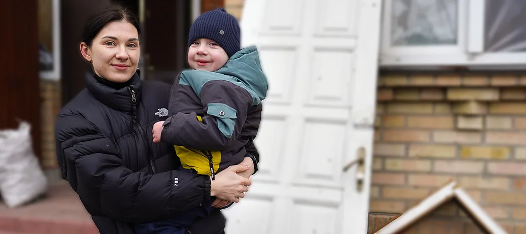 Eine junge Mutter aus der Ukraine hält liebevoll ihren Sohn im Arm, während sie vor einem zerstörten Haus steht, an dessen Wand die Tür gegen die Wand angelehnt ist. Sie benötigen Winterhilfe