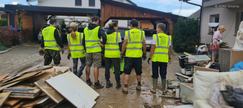 Sechs ADRA-Mitarbeitende mit gelben Westen leisten nach der Hochwasserkatastrophe in Slowenien Hilfe. Im Vorder- und Hintergrund sind durch das Hochwasser zerstörte Möbel und Gegenstände zu sehen