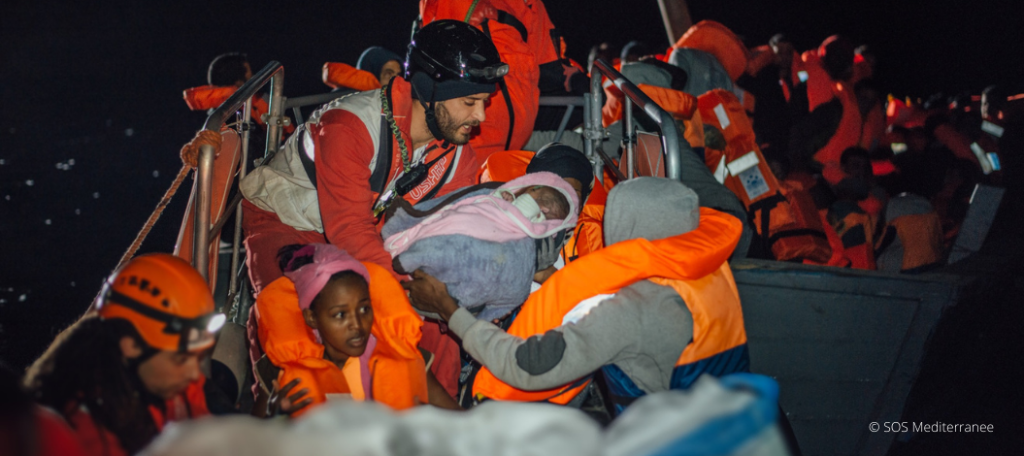Seenotrettung im Mittelmeer durch SOS Mediterranee: Zwei Boote mit geretteten Menschen sind zu sehen. Im Vordergrund überreicht ein Helfer den Eltern ein Kleinkind. Diese bewegende Szene veranschaulicht die lebensrettende Mission der Organisation und die Rettung von Flüchtlingen und Migranten auf See.