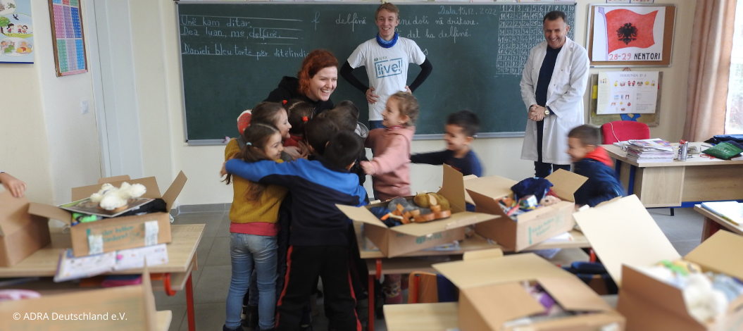 Ein Adralive!-Freiwilliger hilft in einer albanischen Schulklasse bei der Verteilung von Paketen im Rahmen der Aktion 'Kinder helfen Kindern!'. Die Kinder sind aufgeregt und umarmen sich gegenseitig. Im Hintergrund sind die Pakete auf den Schultischen zu sehen.