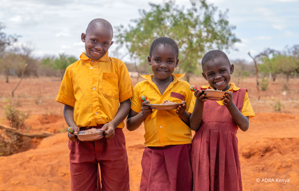 Drei glückliche Schuldkinder aus Kenya in lebhaften rot-gelben Uniformen lachen in die Kamera, während sie einen Teller mit Essen und einen Lutscher in der Hand halten und im Freien stehen