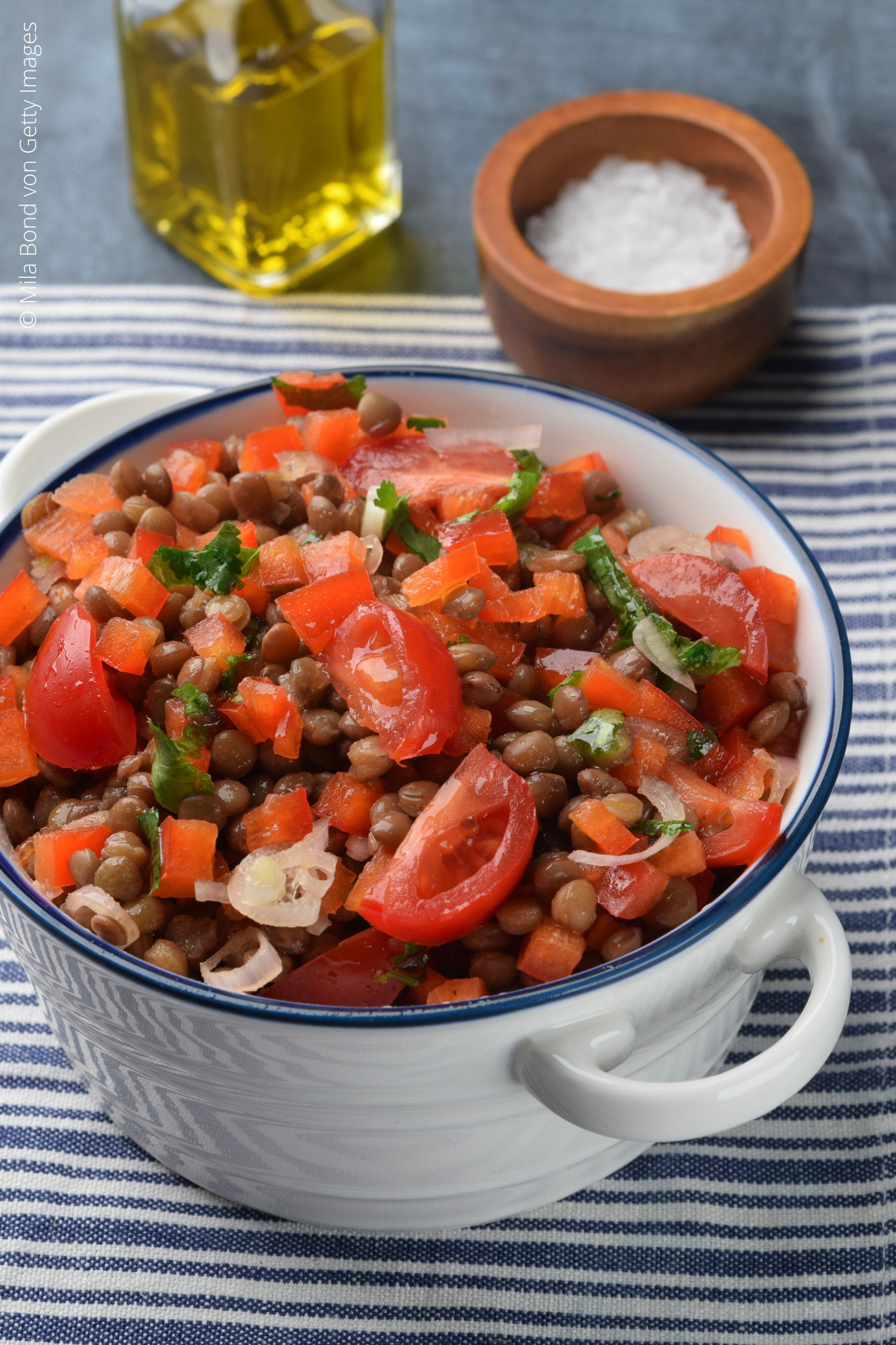Weiße Schüssel mit Linsensalat aus Tomaten, Paprika, Zwiebeln und Kräutern, dahinter Salzschale und Flasche Olivenöl.