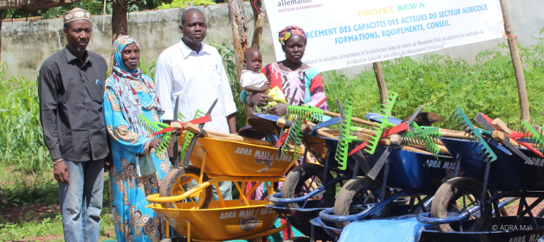 Vier Landwirte und ein Kleinkind in Mali vor Gartenwerkzeugen wie Schubkarren, Gießkannen und Harken