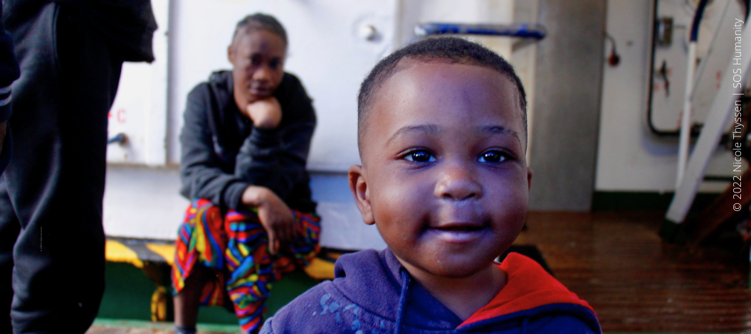 Kleiner Junge blickt zufrieden in die Kamera, Mutter im Hintergrund an Bord des Rettungsschiffs ‚Humanity 1‘ von SOS Humanity