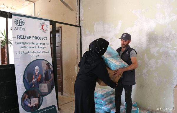 ADRA-Mitarbeiter aus Syrien übergibt einer Frau Hilfspakete nach dem verheerenden Erdbeben. Im Hintergrund ein ADRA-Banner mit den Worten 'Hilfsprojekt - Notfallmaßnahmen nach dem Erdbeben in Aleppo'.