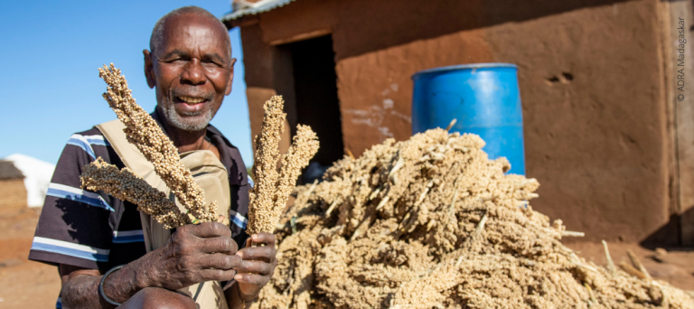 Lächelnder Mann aus Madagaskar sitzt im Freien und hält Getreide, hinter ihm ein großer Berg voller Getreide.