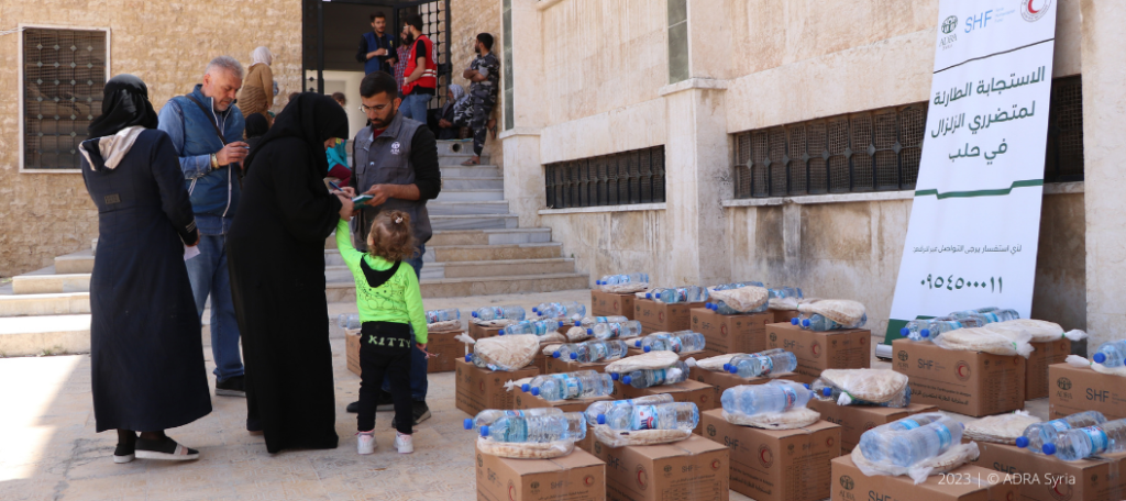 Bereitgestellte Lebensmittelpakete vor einem ADRA-Roll-Up-Plakat für die Erdbebenhilfe in Syrien, während Menschen auf humanitäre Unterstützung warten.