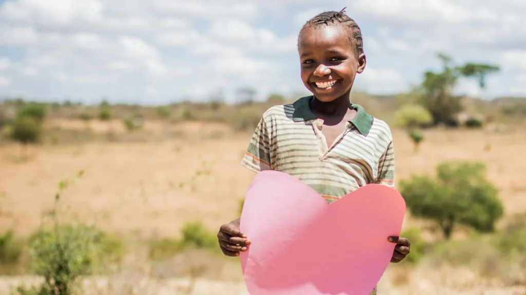 Ein strahlendes Kind in Kenia hält ein großes rotes Papierherz mit beiden Händen vor sich. Ein Bild voller Mitgefühl, Gerechtigkeit und Liebe, während es direkt in die Kamera lächelt.