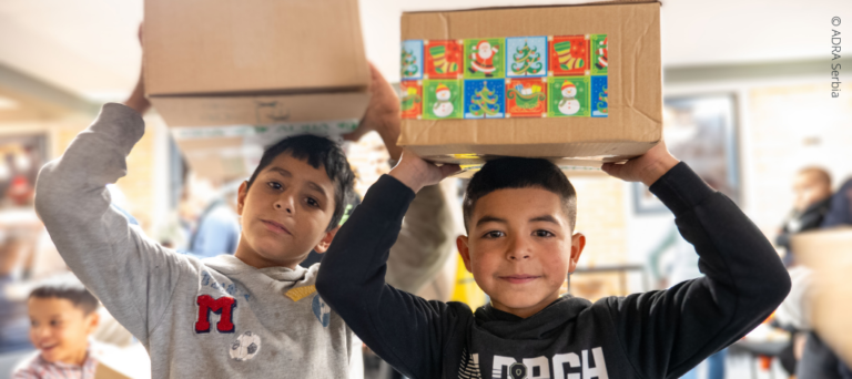 Zwei Kinder in Serbien heben strahlend ihre Pakete der ‚Aktion Kinder helfen Kindern!‘ über die Köpfe