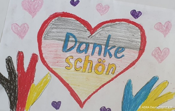 Eine Kinderzeichnung zeigt ein großes rotes Herz, das mit der deutschen Flagge gefüllt ist und das Wort 'Dankeschön' trägt. Unterhalb des Herzens sind zwei Hände dargestellt, eine in den Farben der deutschen Flagge und die andere in den Farben der Ukraine