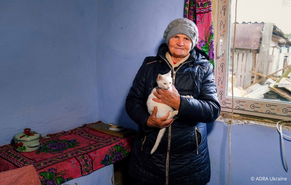 Eine ältere Dame namens Irina aus der Region Mykolaiv steht in ihrer Wohnung. Sie trägt eine Winterjacke, Mütze und Schal und hält eine Katze im Arm. Hinter ihr ist ein frisch eingebautes neues Fenster zu sehen.