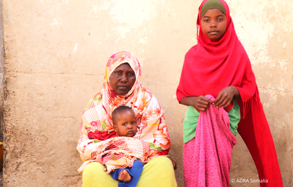 Mutter sitzt vor einer Mauer und hält ihr Kleinkind im Schoß, daneben steht ein weiteres Kind