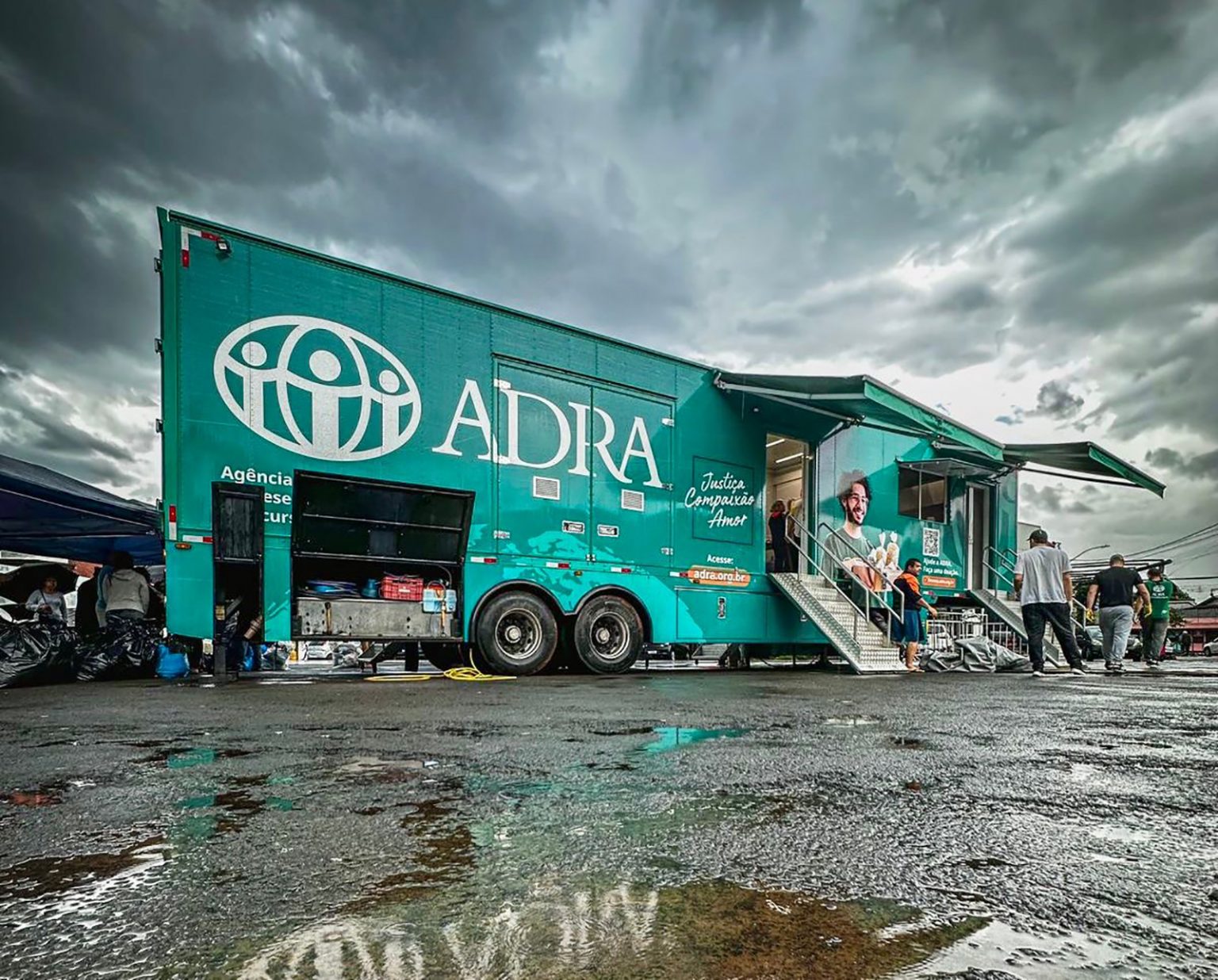 Grüner ADRA-Hilfslastwagen in Brasilien, bei nassem Wetter im Einsatz. Helferinnen und Helfer bewegen sich rund um das Fahrzeug, auf dessen Seite das ADRA-Logo deutlich sichtbar ist.