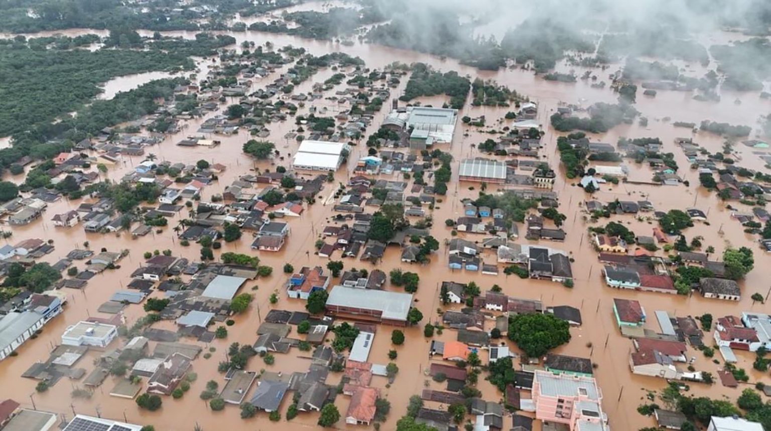Luftaufnahme eines überfluteten Stadtgebiets in Brasilien. Zahlreiche Häuser und Straßen stehen im braunen Hochwasser, nur Dächer und einige höhere Gebäude ragen heraus.
