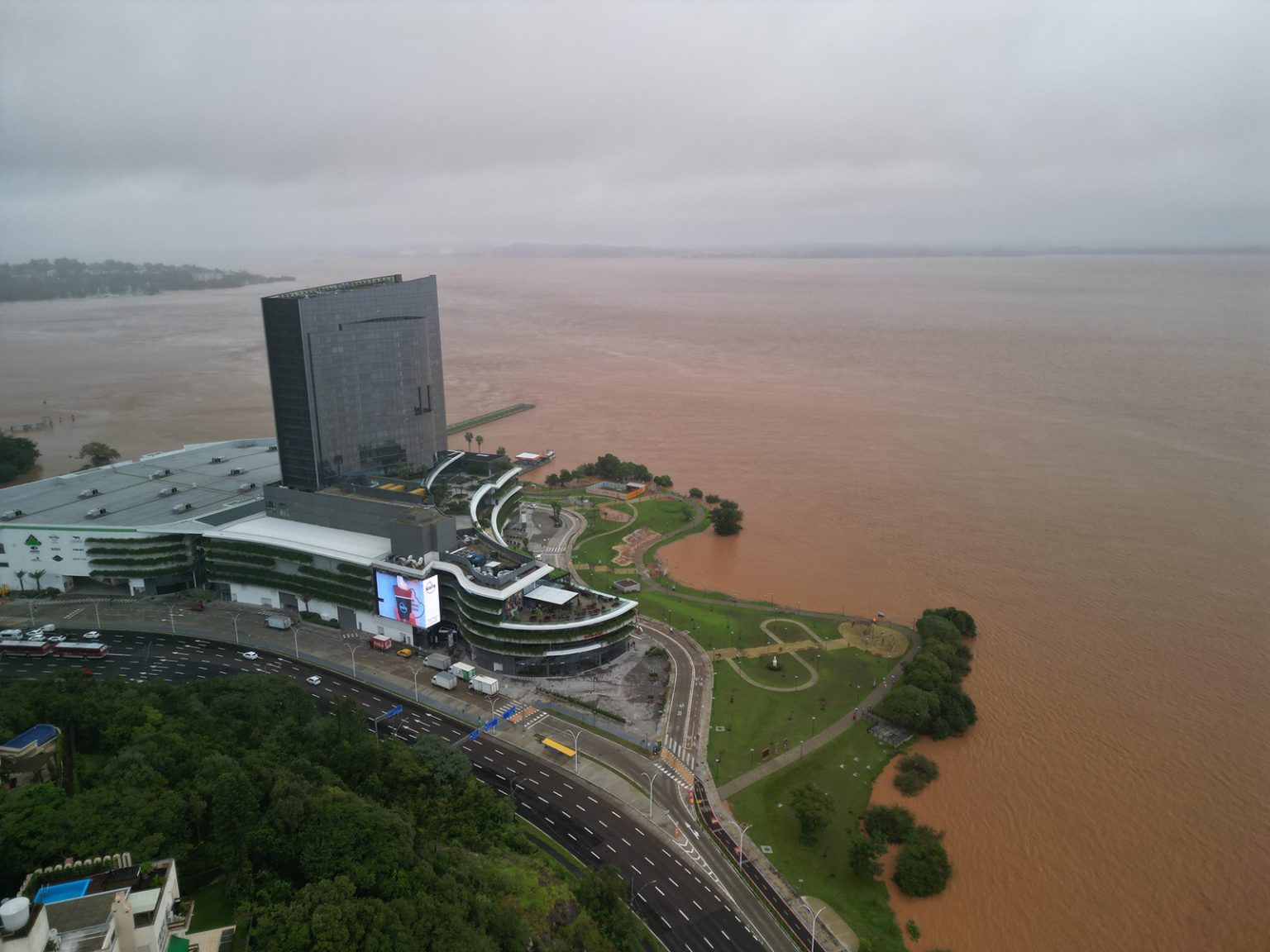 Modernes Gebäude an einem Flussufer in Brasilien, das vom stark angestiegenen, braunen Hochwasser umgeben ist. Uferbereiche und Grünflächen sind teilweise überflutet.