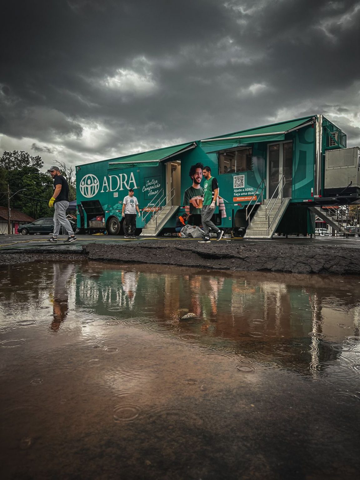 Seitenansicht eines grünen ADRA-Hilfslastwagens aus Brasilien mit ausgeklappten Treppen und Markisen. Mehrere Personen stehen davor, der Himmel ist stark bewölkt.
