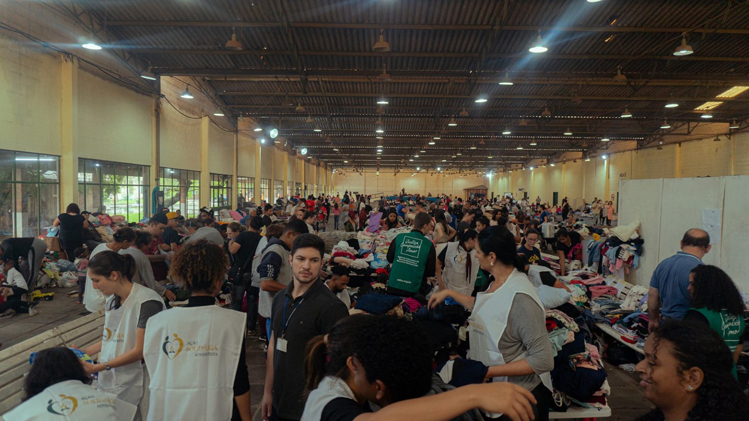 Große Halle in Brasilien mit zahlreichen freiwilligen Helferinnen und Helfern, die gespendete Kleidung sortieren und verteilen. Tische sind mit Kleidung bedeckt, viele Menschen arbeiten gleichzeitig im Raum.
