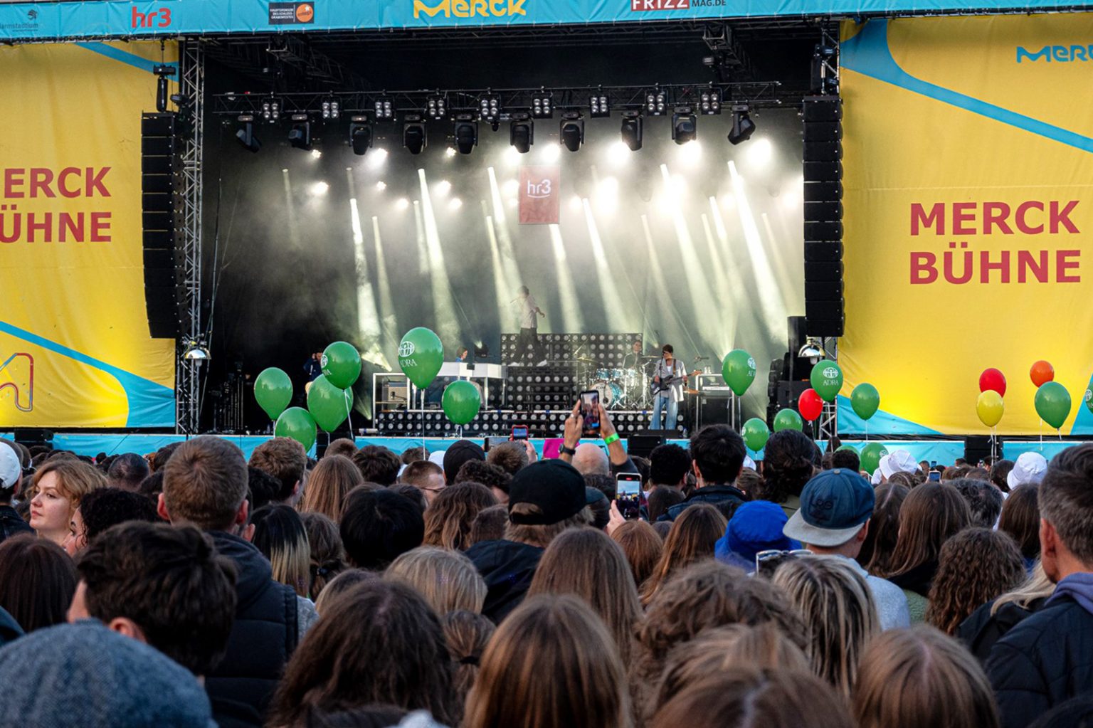 Große Konzertbühne mit Lichtshow und dichtem Publikum. Mehrere grüne ADRA-Luftballons ragen aus der Menge hervor.