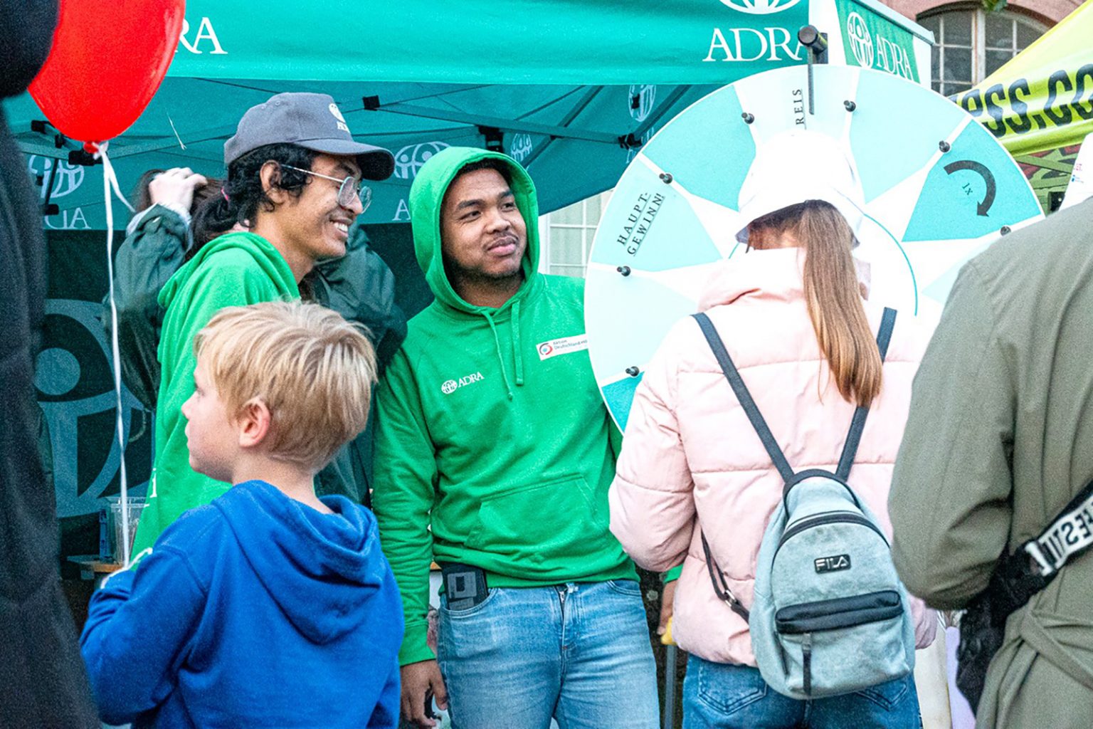 Zwei ADRA-Mitarbeitende in grünen ADRA-Hoodies sprechen mit Besucherinnen und Besuchern vor einem Glücksrad unter einem ADRA-Pavillon.