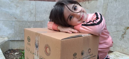 Ein junges Mädchen aus Syrien lächelt in die Kamera, während sie ihren Kopf auf ein Hilfsgüter-Paket gelegt hat.