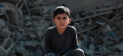 Ein Junge sitzt mit ernstem Blick vor zerstörten Gebäudetrümmern, Symbol für die Not und Unsicherheit in Krisengebieten