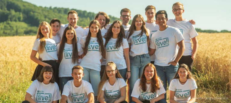 Gruppenfoto von ADRAlive!-Freiwilligen auf einem Feld: Eine Gruppe von Freiwilligen steht lächelnd auf einem offenen Feld, umgeben von grüner Landschaft. Sie tragen ein weißes T-Shirt mit dem Logo von ADRAlive! und sind in guter Stimmung.