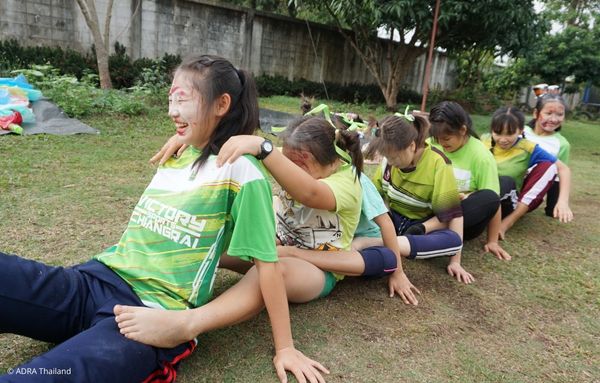 Kinder und Jugendliche aus Thailand spielen lachend ein Gruppenspiel im Freien, bei dem sie hintereinander auf dem Boden sitzen und sich an den Schultern halten.