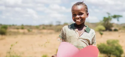 Lächelndes Kind in Kenia hält ein großes rotes Papierherz mit beiden Händen vor sich.