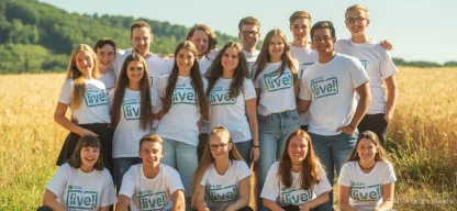 Gruppenfoto von ADRAlive!-Freiwilligen auf einem Feld: Eine Gruppe von Freiwilligen steht lächelnd auf einem offenen Feld, umgeben von grüner Landschaft. Sie tragen ein weißes T-Shirt mit dem Logo von ADRAlive! und sind in guter Stimmung.
