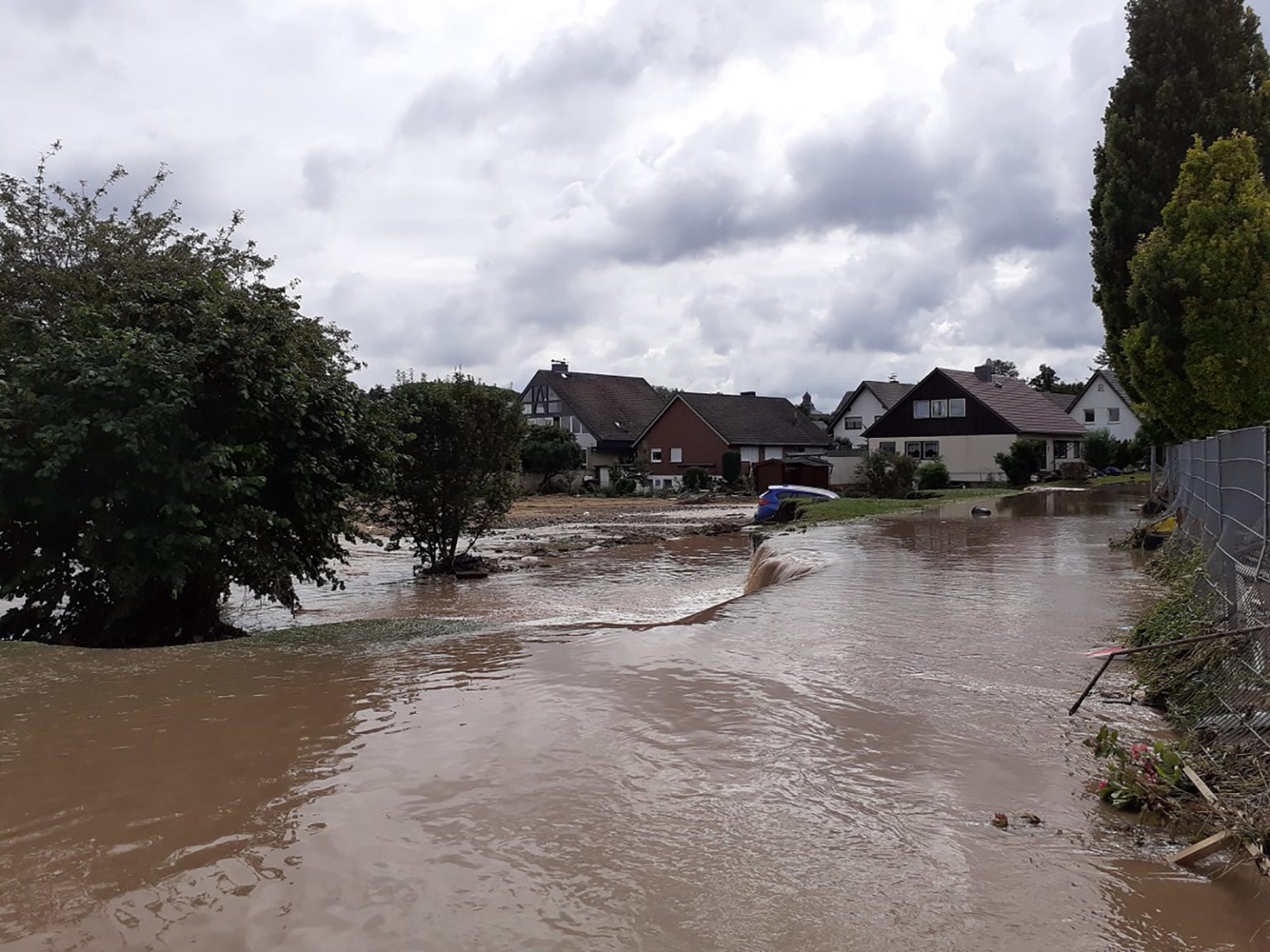 Dieses Bild zeigt, wie die Wassermassen der Erft über den Sportplatz und durch den Zaun ins benachbarte Feld fließen. Einen Teil der Anliegerstraße hat die Flut weggerissen. (Foto: RSV Arloff-Kirspenich e.V.)