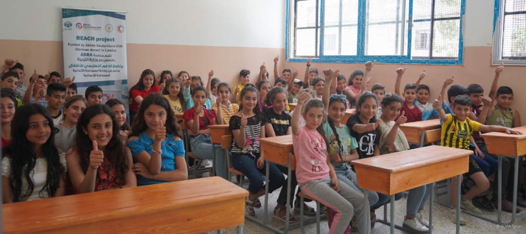 Eine gemischt Schulklasse sitzt in einem Klassenzimmer. Alle Kinder strecken die Hände nach oben und strahlen Freude aus.