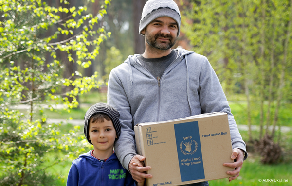 Ein Mann steht mit einem Jungen und einem Lebensmittelpaket vor Bäumen