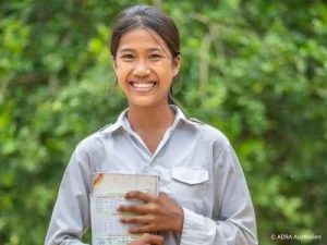 Junge Frau steht draußen vor grünem Hintergrund, lächelt und hält Schulhefte im Arm.