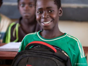 Ein Junge sitzt in einem Klassenzimmer und hält glücklich seinen Schulranzen auf dem Schoß.