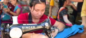 Eine Frau in Thailand sitzt an einem Tisch und näht konzentriert an einer Nähmaschine an einem Kleidungsstück.