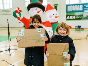 Zwei Kinder aus Osteuropa stehen in einer Turnhalle und halten ein Paket der Aktion „Kinder helfen Kindern!“ in den Händen. Beide strahlen vor Freude über ihr Weihnachtsgeschenk.