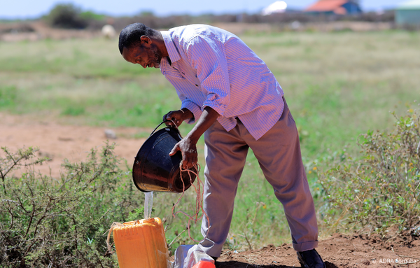 Mann aus Somalia steht in einem Feld und schüttet Wasser aus einer Schüssel in einen Kanister – Symbol für den Alltag und die Herausforderungen in ländlichen Regionen