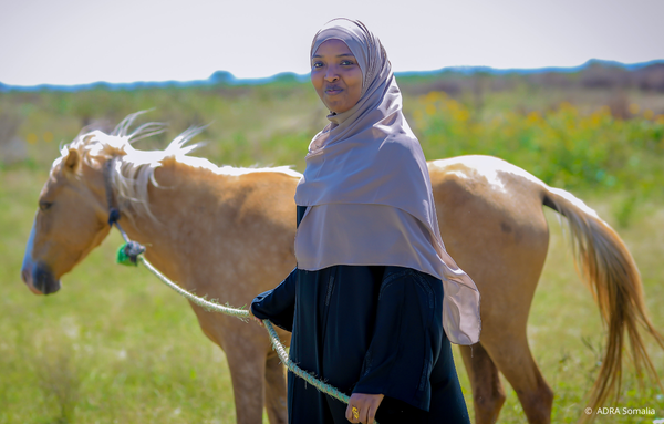 Frau in Somalia mit Kopftuch führt ein Pferd an der Leine und grinst in die Kamera