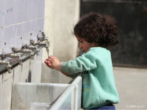 Ein Kind wäscht sich die Hände an einem langen Waschbecken mit mehreren Wasserhähnen. Das Bild symbolisiert den Zugang zu sauberem Trinkwasser und die Wiederherstellung von Wasserversorgungsanlagen durch ADRA-Projekte in Konfliktgebieten.