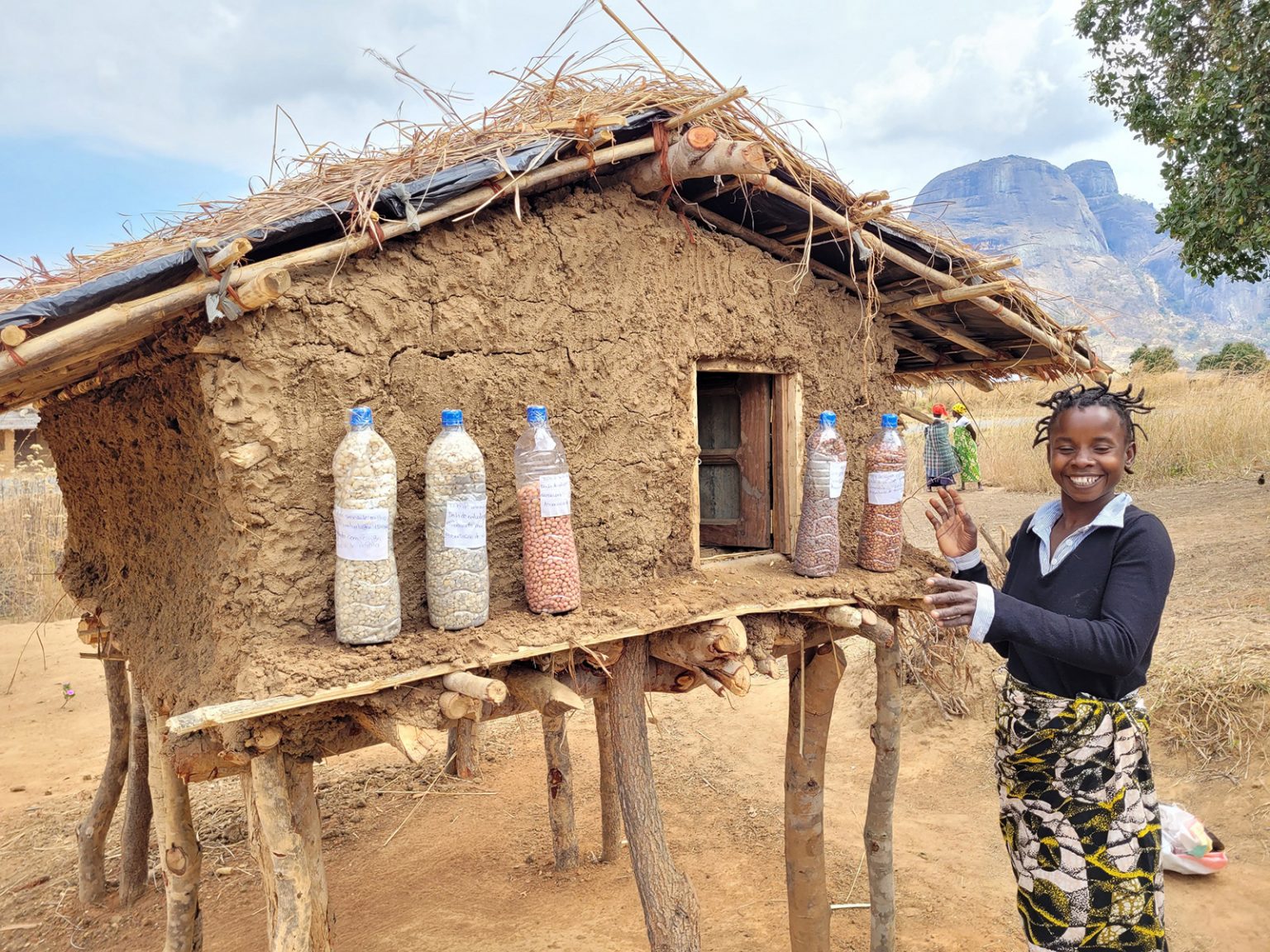 Ein Mädchen steht vor einer Saatgutbank in Mosambik, die mit Plastikflaschen voller Saatgut gefüllt ist. Sie lächelt stolz, während sie die verschiedenen Saatgutsorten präsentiert, die für nachhaltige Landwirtschaft und Klimaanpassung in der Region von entscheidender Bedeutung sind.