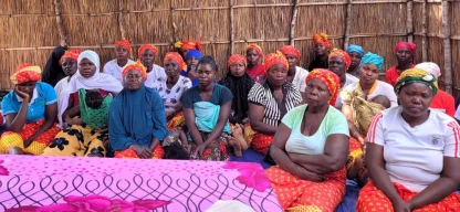 Eine Gruppe von Frauen und Kindern aus Mosambik sitzt auf dem Boden und hört aufmerksam zu. Das Bild ist lebendig und bunt, was die Energie und den Gemeinschaftssinn der Gruppe widerspiegelt.