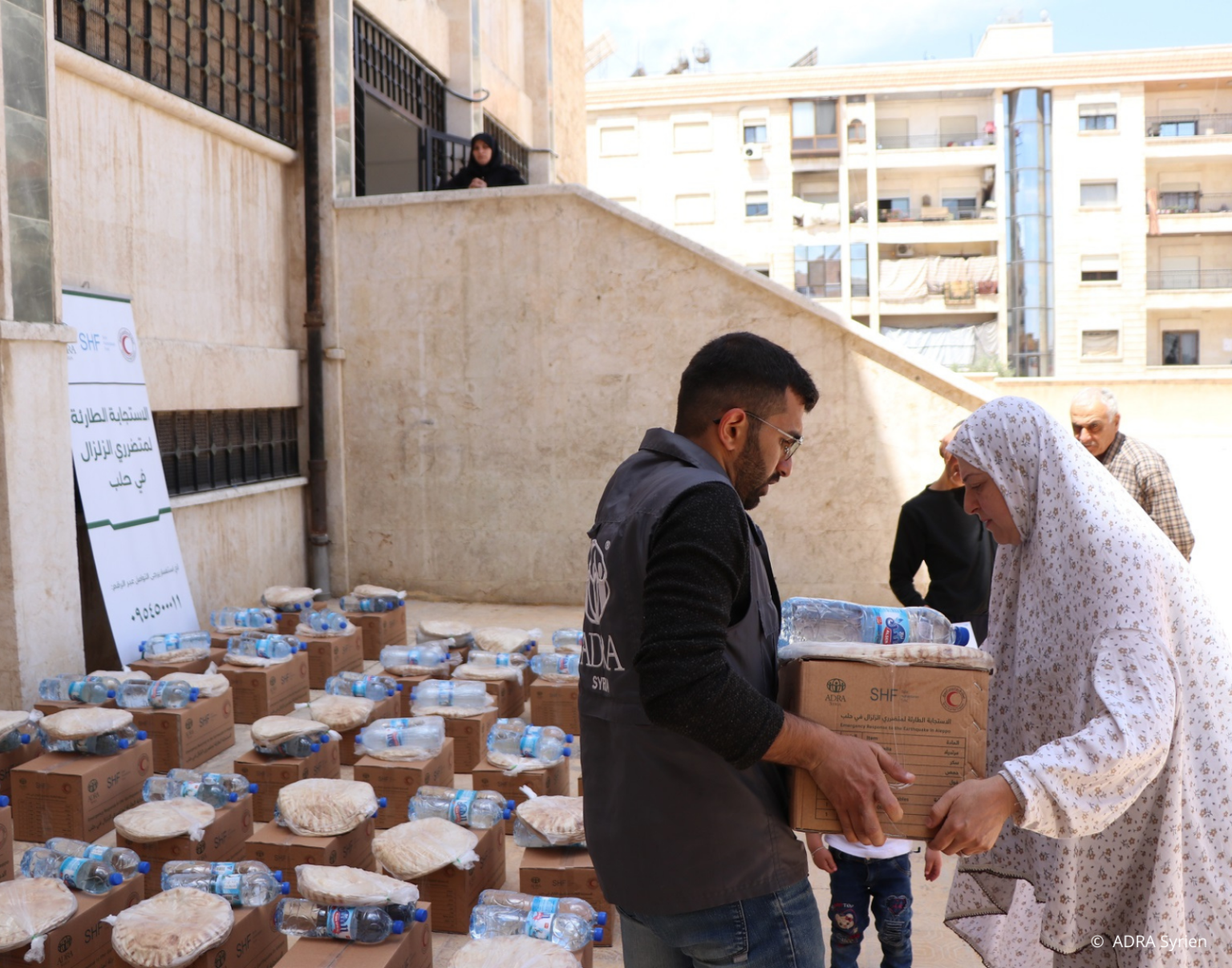 ADRA-Helfer überreicht syrischer Frau Hilfspaket mit Wasser und Fladenbrot – umgeben von weiteren gepackten Kartons