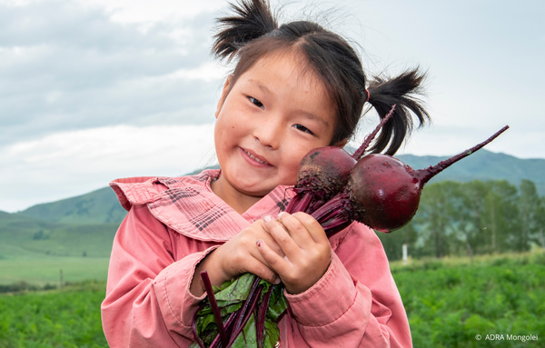 Ein kleines Mädchen aus der Mongolei steht auf einem weiten Feld, hält frisch geerntete rote Beete in den Händen und lächelt glücklich in die Kamera. Sie freut sich über den erfolgreichen Ertrag