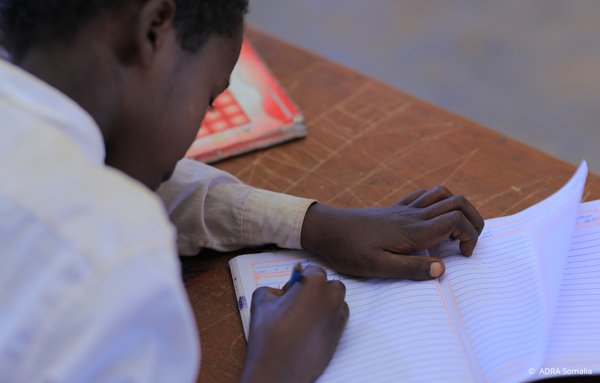 Ein somalischer Junge in Schuluniform konzentriert sich auf das Schreiben in seinem Heft, ohne in die Kamera zu schauen. Das Bild zeigt, wie Bildungsinitiativen von ADRA und Save the Children Kindern im Krisengebiet Somalia helfen, wieder am Unterricht teilzunehmen