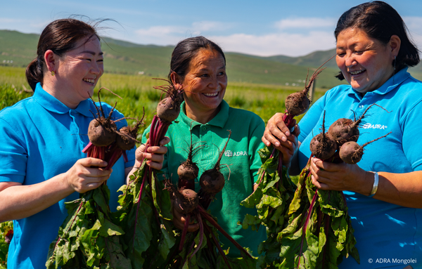 Drei Frauen aus der Mongolei stehen auf einem weiten Feld, halten frisch geerntete rote Beete in den Händen und lachen fröhlich in die Kamera. Sie freuen sich über den reichen Ertrag ihrer Bio-Landwirtschaft